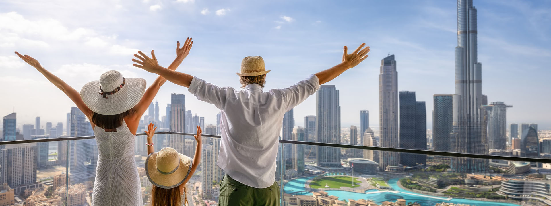 Three people in hats stand on a balcony with arms raised, overlooking a modern city skyline with tall skyscrapers on a sunny day.