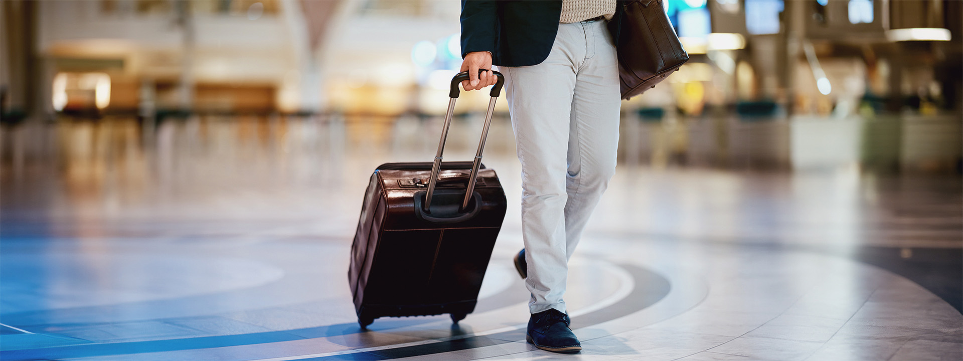 A person in a suit jacket, white pants, and dark shoes walks through an airport with a wheeled suitcase and a shoulder bag.