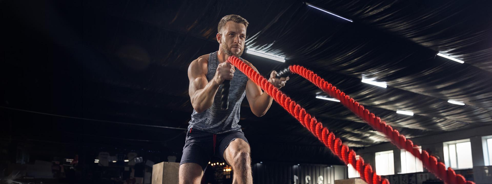 Man in a gym working out with red battle ropes, wearing a gray tank top and black shorts, with a focused expression.