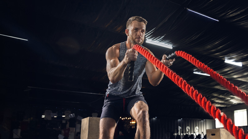 Man in a gym working out with red battle ropes, wearing a gray tank top and black shorts, with a focused expression.