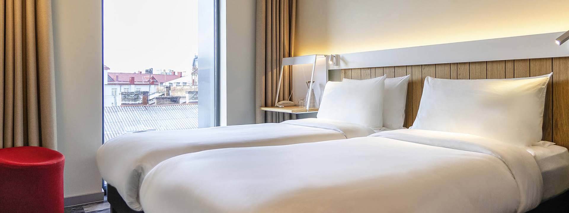 A modern hotel room with two single beds, white bedding, wooden headboard, bedside table with lamp, large window, beige curtains, and a red round stool.