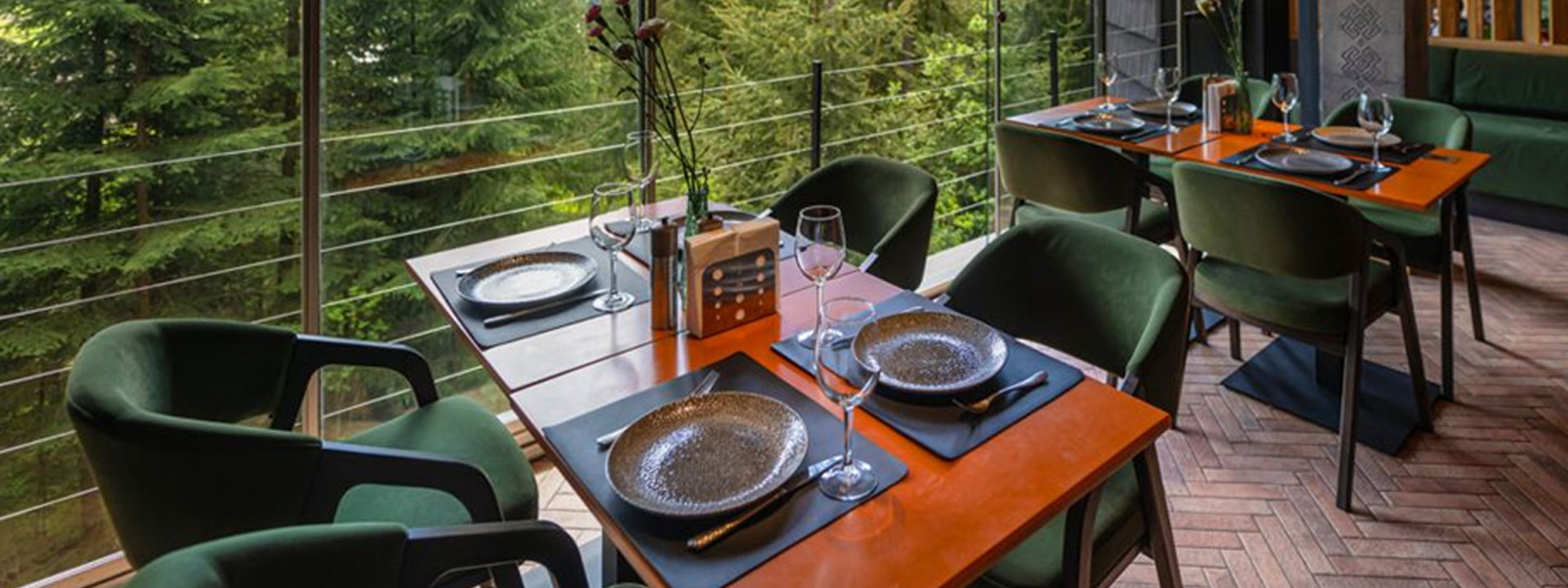 A modern restaurant dining area with dark green chairs, orange tables set with plates and glasses, and large windows overlooking trees.