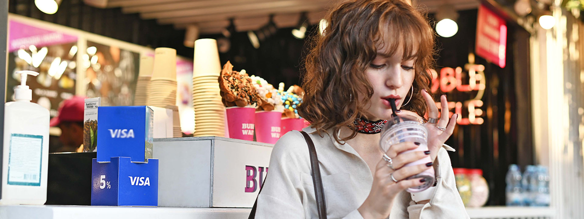 A woman enjoys a drink through a straw at a food stall decorated with visa branding and hand sanitizer visible.