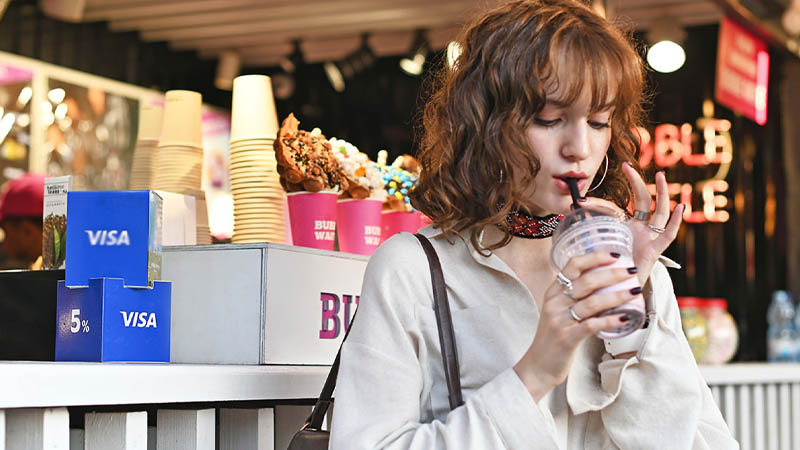 A woman enjoys a drink through a straw at a food stall decorated with visa branding and hand sanitizer visible.