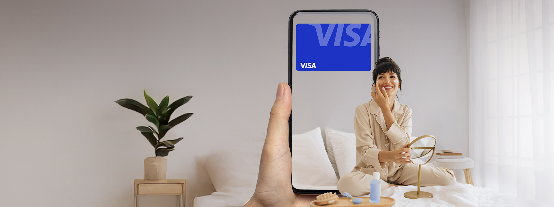 A person in beige pajamas sits on a bed and smiles while holding a phone that displays a large blue Visa logo in a bright, minimalist bedroom.