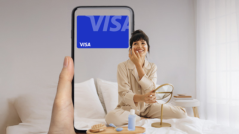 A person in beige pajamas sits on a bed and smiles while holding a phone that displays a large blue Visa logo in a bright, minimalist bedroom.