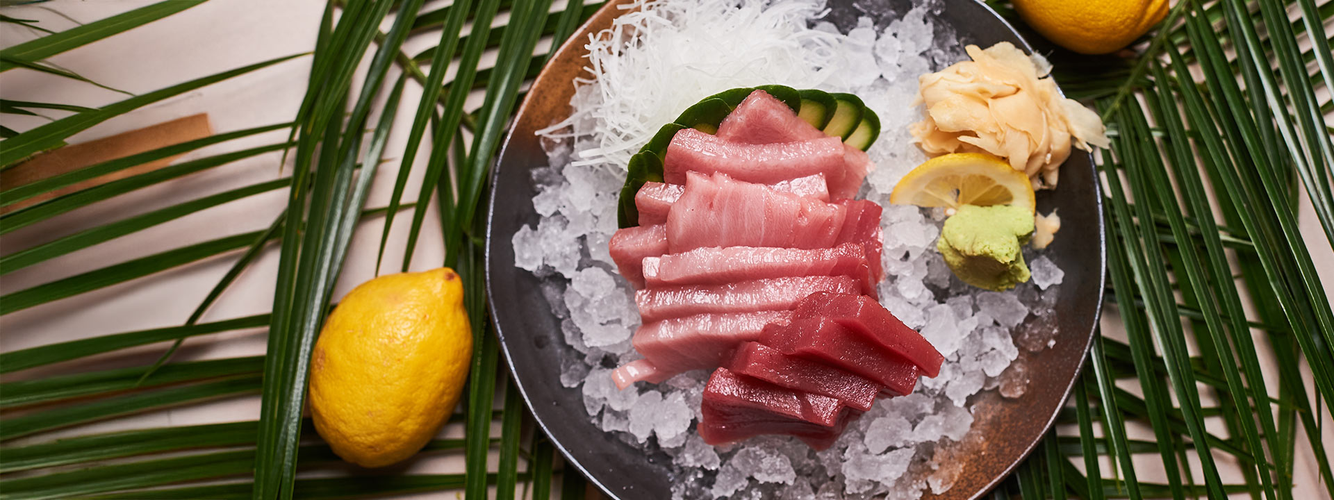 A plate of sashimi slices on ice with shredded daikon, cucumber, pickled ginger, wasabi, a lemon slice, and two whole lemons on a palm leaf background.