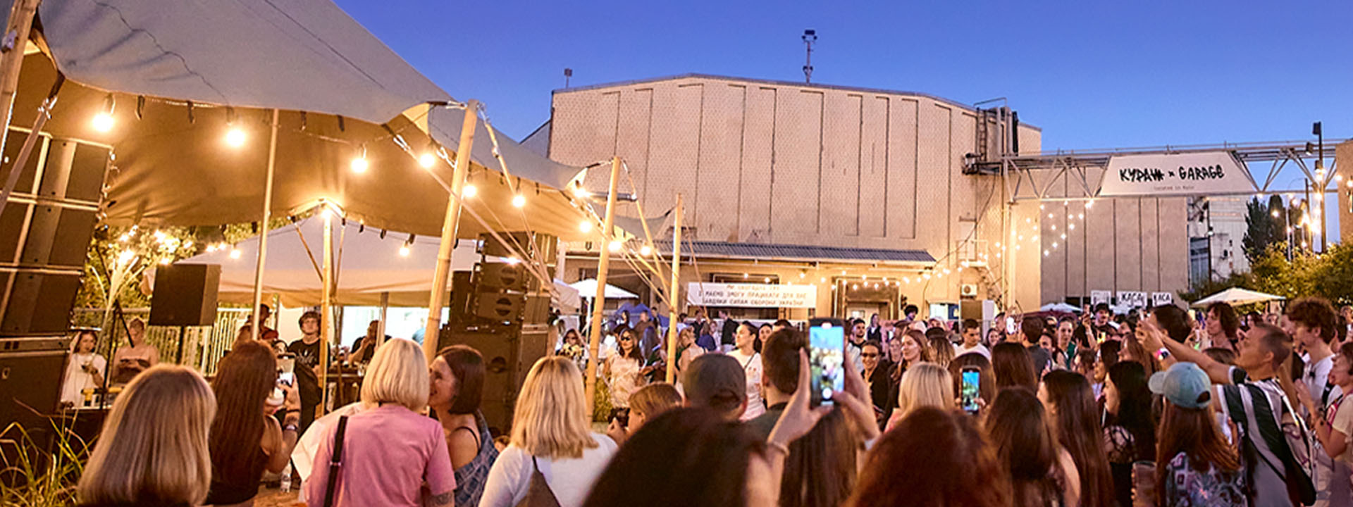 A crowd of people gathers outdoors under string lights at a lively event during twilight.