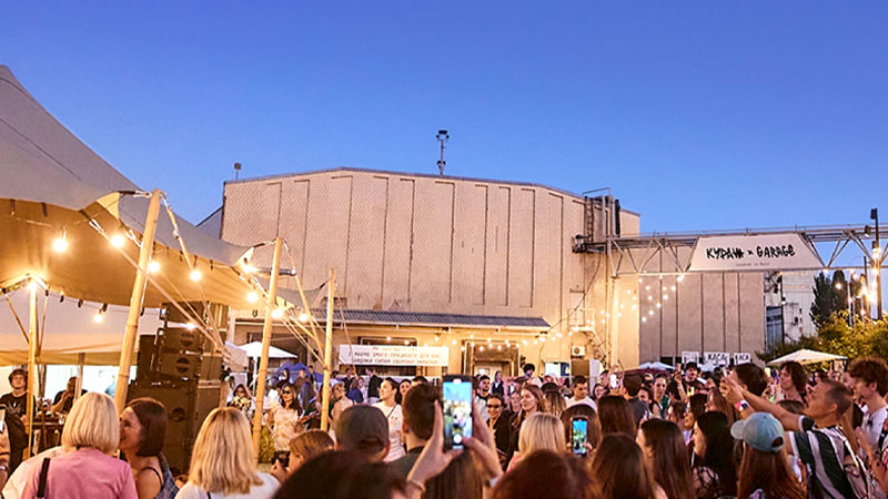 A crowd of people gathers outdoors under string lights at a lively event during twilight.