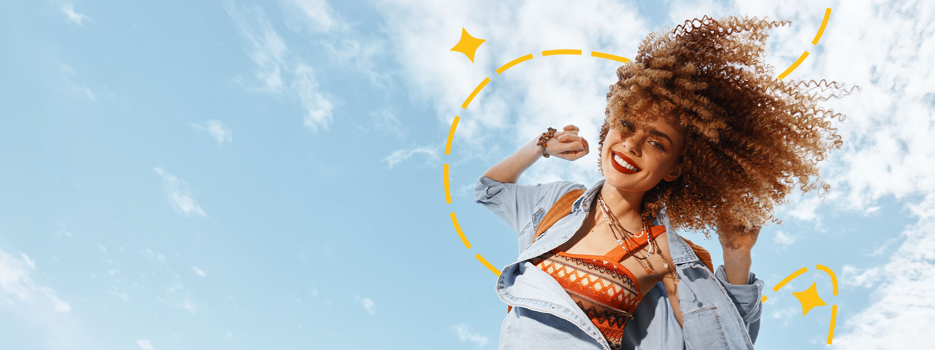 A woman with curly hair smiles and poses outdoors against a blue sky, wearing a patterned top and denim jacket, with yellow graphic accents around her.