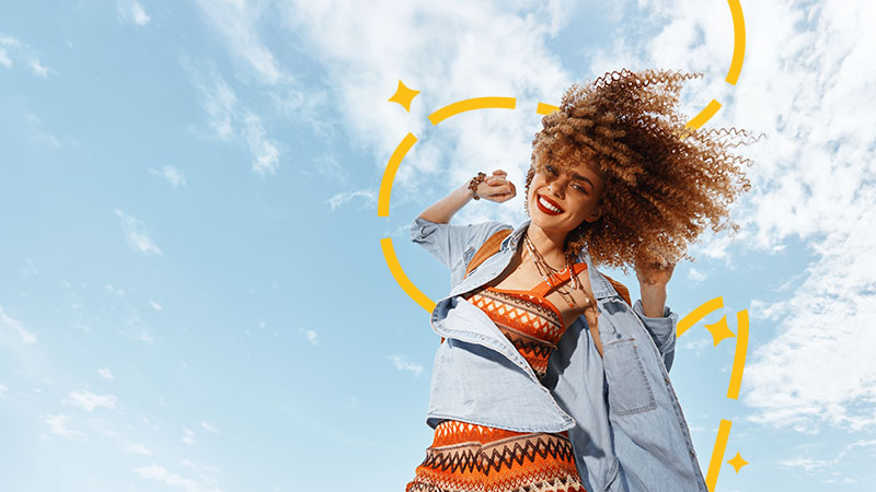 A woman with curly hair smiles and poses outdoors against a blue sky, wearing a patterned top and denim jacket, with yellow graphic accents around her.