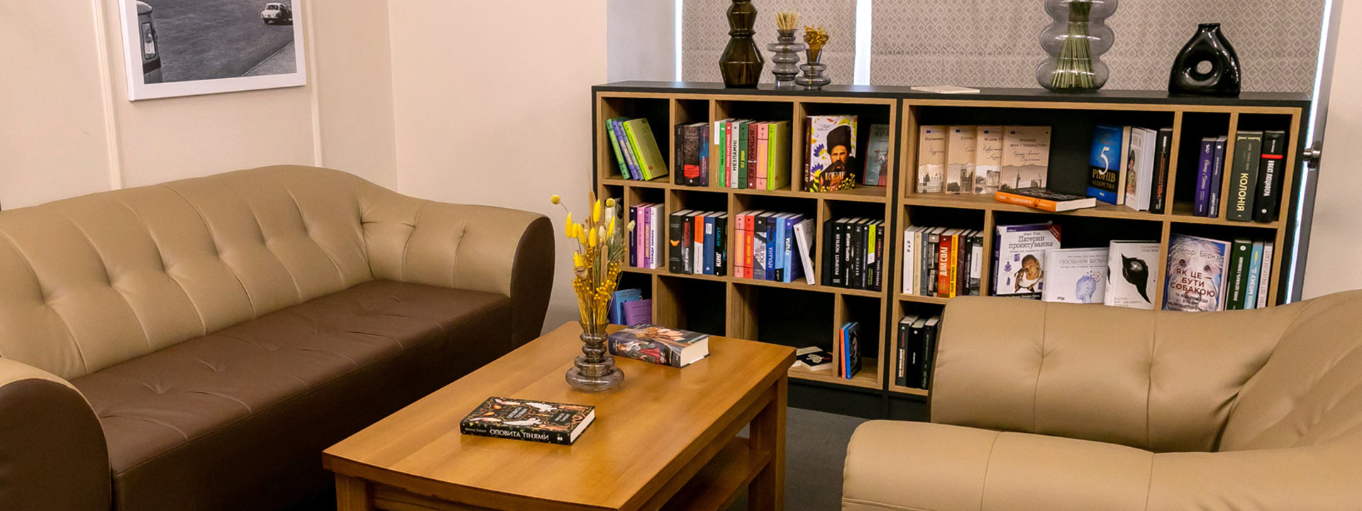 A cozy reading area with two beige sofas, a wooden coffee table holding books and vases, and a bookshelf filled with books and decorative items in the background.