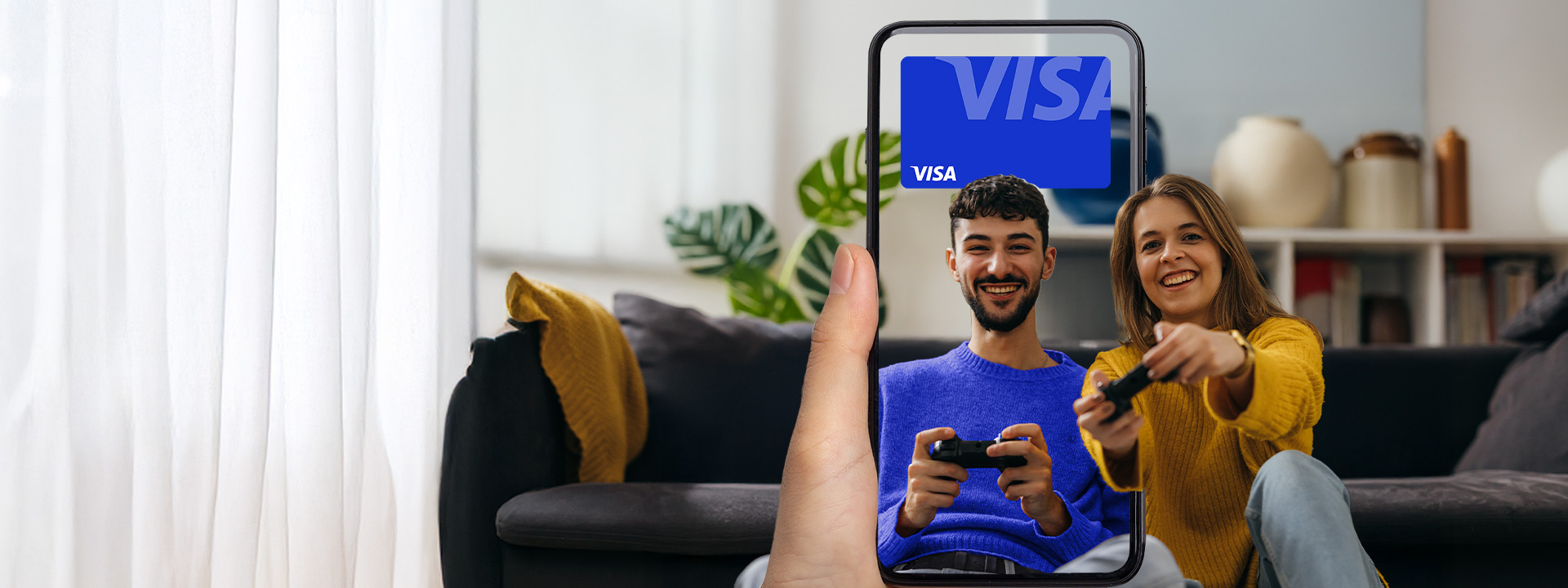 A hand holds a phone displaying a digital Visa card; in the background, a smiling man and woman sit on a couch playing video games.