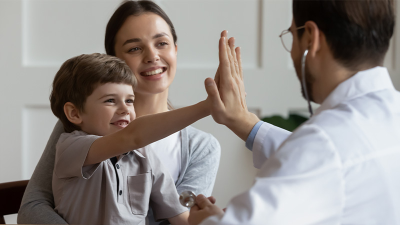 A child gives a high-five to a person in a white coat while a smiling woman watches.