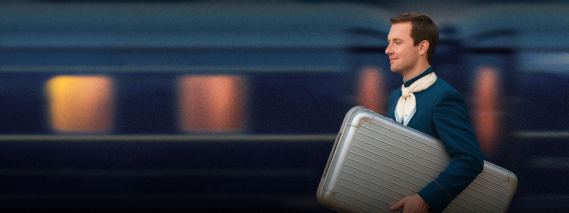 A man in a blue uniform and neck scarf walks in front of a blurred moving train, carrying a large silver suitcase.