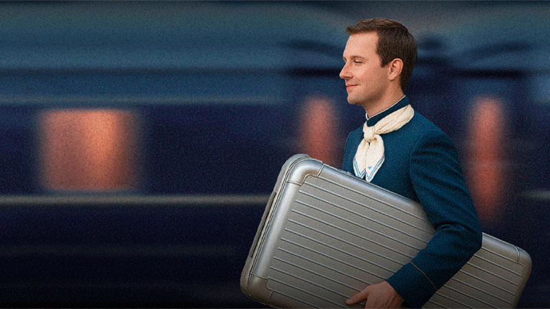 A man in a blue uniform and neck scarf walks in front of a blurred moving train, carrying a large silver suitcase.