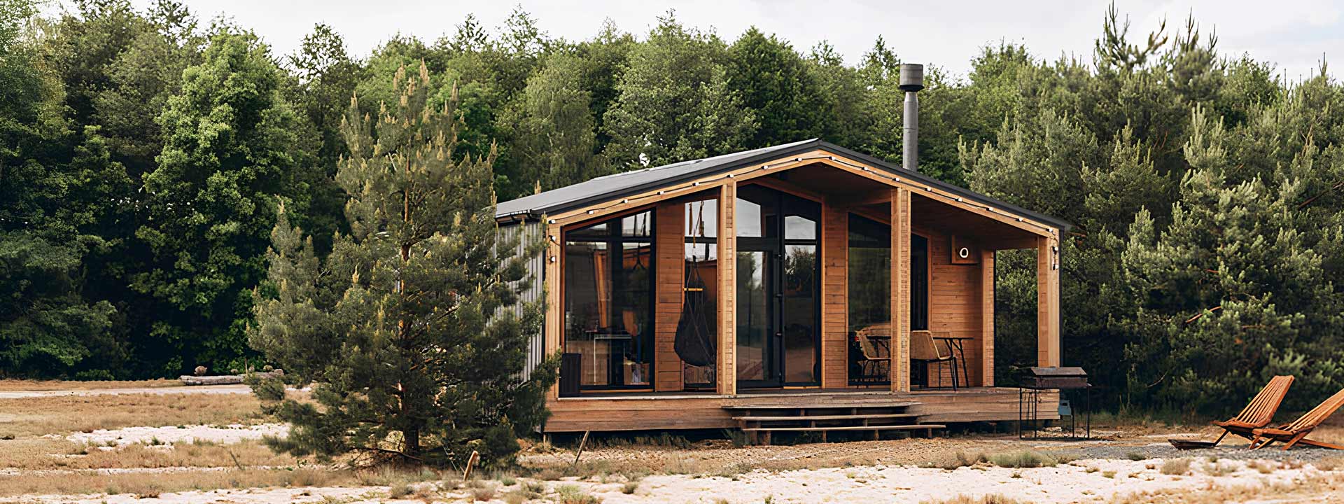 A small, modern wooden cabin with large windows, surrounded by trees, sits in a clearing with a hammock and a sun chair on a porch.