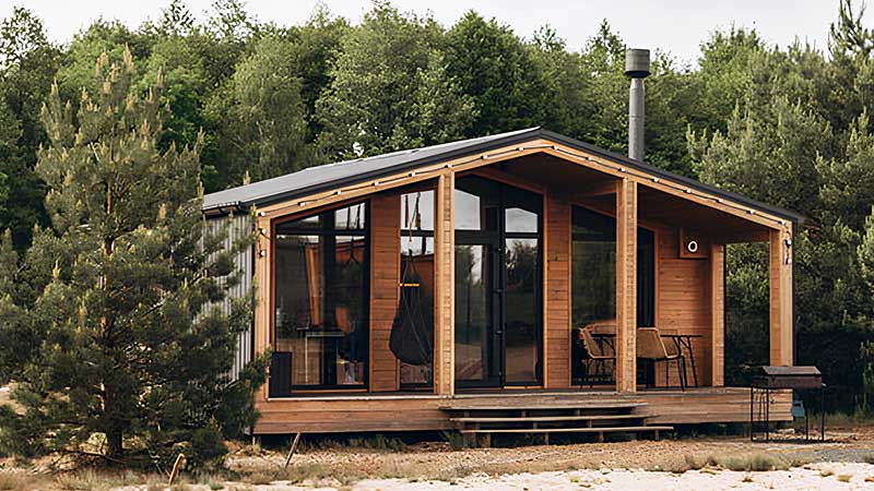 A small, modern wooden cabin with large windows, surrounded by trees, sits in a clearing with a hammock and a sun chair on a porch.