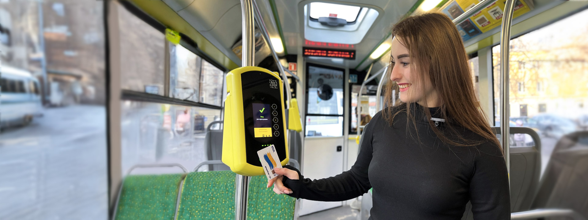 A person with long hair, wearing a black top, uses a card on a yellow ticket machine inside a public bus.