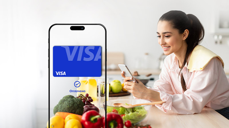 A person sitting at a white table filled with fruits and vegetables is holding a smartphone with a Visa advertisement displayed on the screen, symbolizing a digital transaction or online payment.