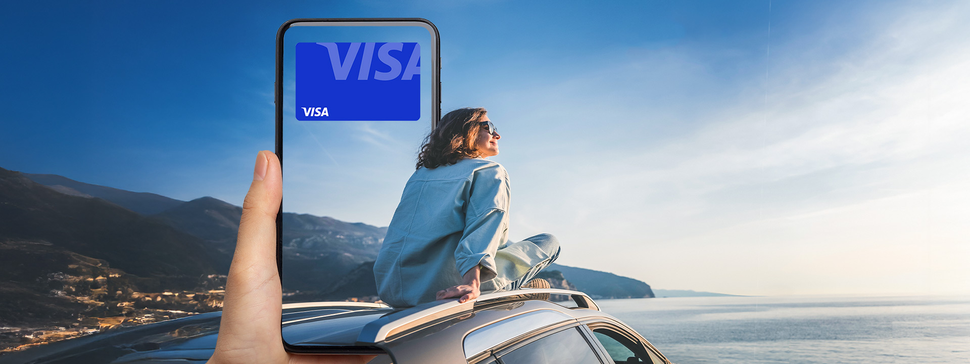 A person sits on top of a car by the ocean, with a hand holding a phone in the foreground displaying a digital Visa card.