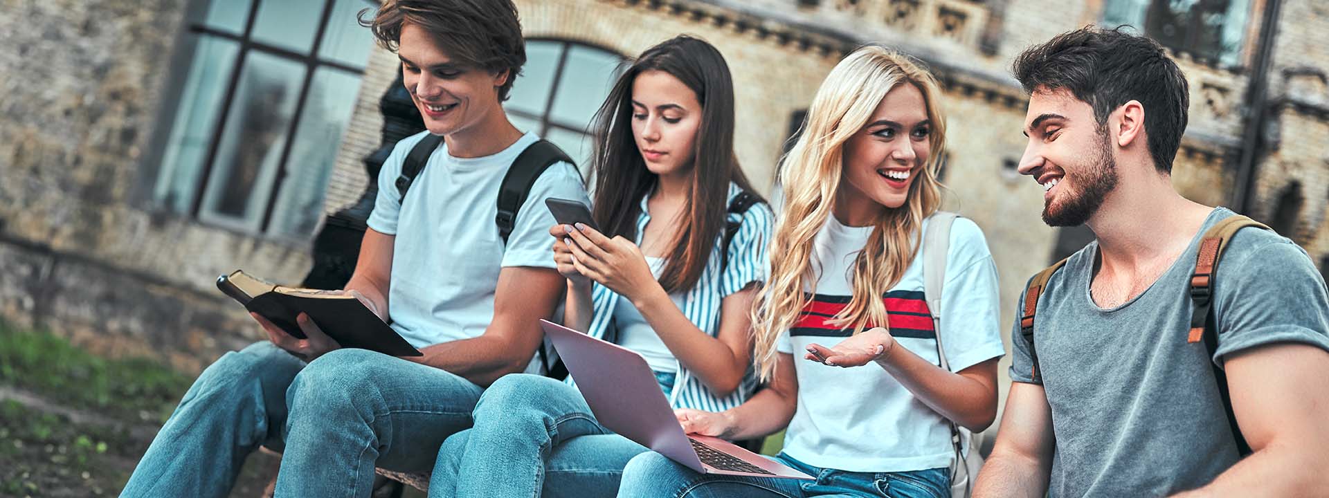 Four students sitting outdoors, with one reading a book, another using a smartphone, a third looking at a laptop, and all casually interacting.