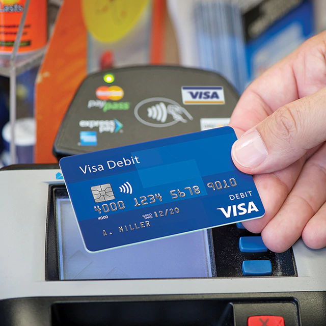 Person holding a Visa credit card in front of a payment terminal displaying the contactless symbol and credit card logos.