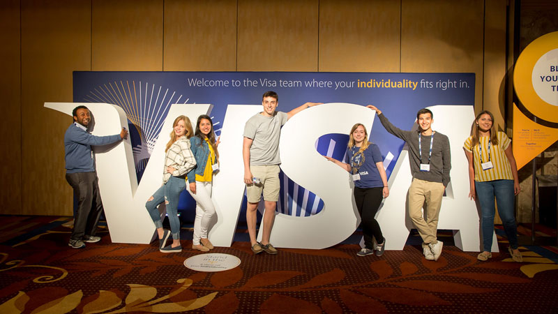 A co-ed group of smiling interns pose in front of a big Visa sign.