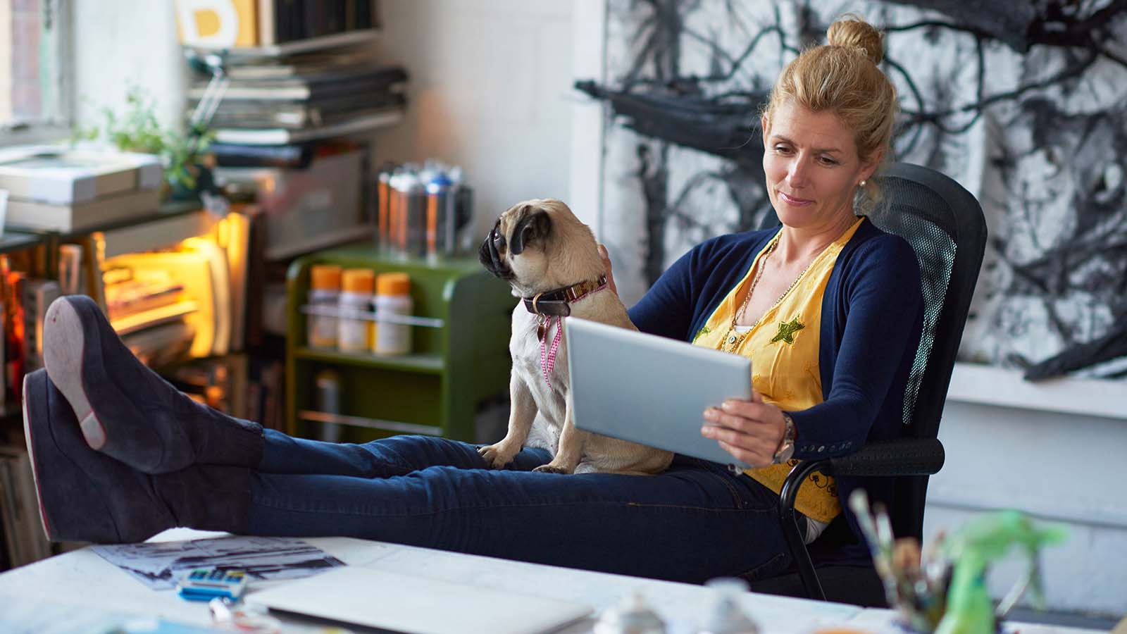 Woman with her feet on a desk and a dog in her lap reading a tablet. 