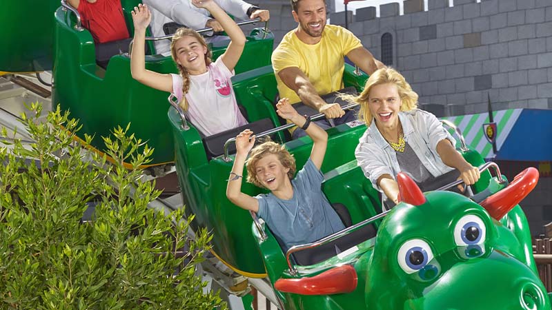 Four people, two adults and two children, ride a green dragon-themed roller coaster, all smiling and raising their hands. The background includes greenery and a castle-like structure.