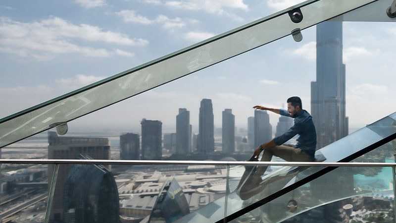 A man sits on a glass staircase, elevated above a city skyline with several modern skyscrapers visible in the background on a partly cloudy day.