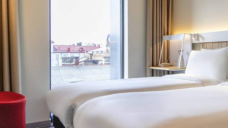 A modern hotel room with two single beds, white bedding, wooden headboard, bedside table with lamp, large window, beige curtains, and a red round stool.