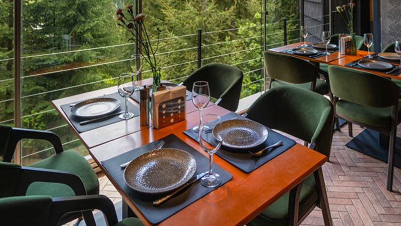 A modern restaurant dining area with dark green chairs, orange tables set with plates and glasses, and large windows overlooking trees.
