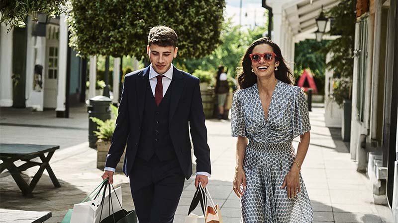 A man in a suit and a woman in a patterned dress with sunglasses walk outdoors holding shopping bags.