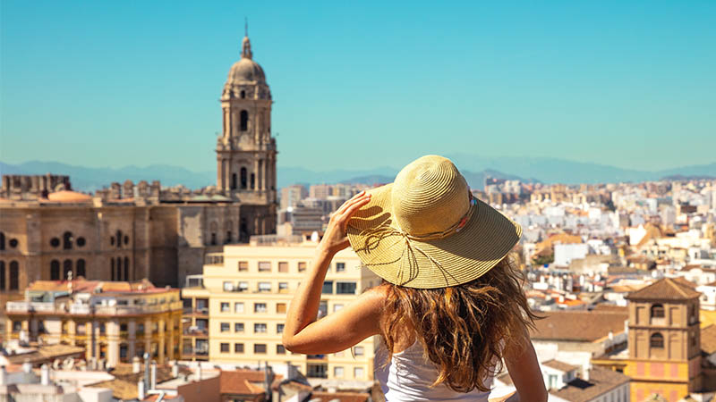 A woman in a straw hat overlooks a city's scenic skyline with a prominent historic cathedral under a clear blue sky.
