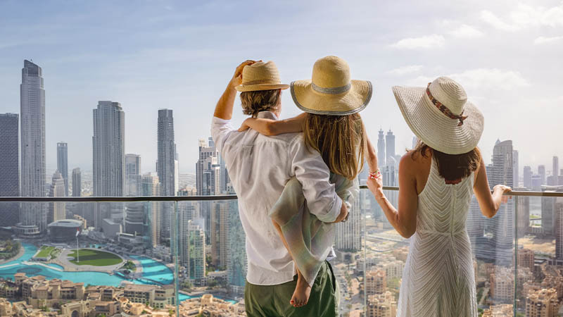 A family of three wearing hats stands on a balcony overlooking a city with tall buildings and a distant view of a body of water.