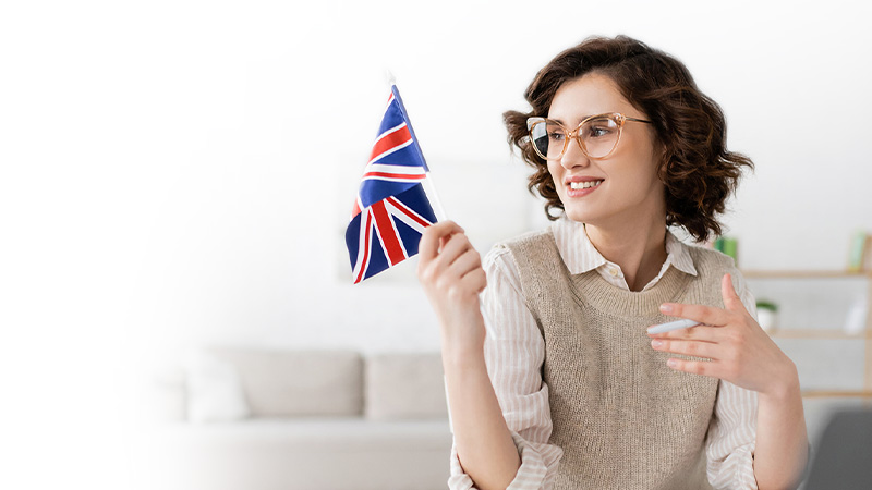 A smiling young woman with curly hair and glasses holding and looking at a small union jack flag in an indoor setting.