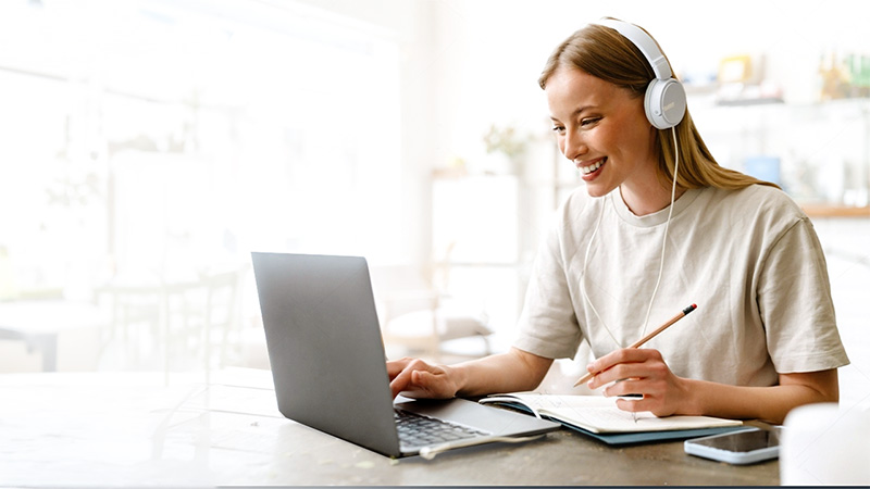 A young woman wearing headphones smiles while using a laptop and taking notes at a bright cafe table.