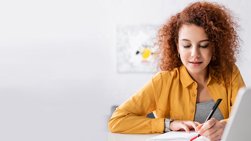 A focused young woman with curly red hair, wearing a yellow shirt, writing in a notebook at a white table.