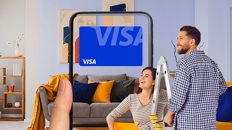 A person holds a smartphone displaying a virtual Visa card in front of a couple painting and decorating a modern living room.