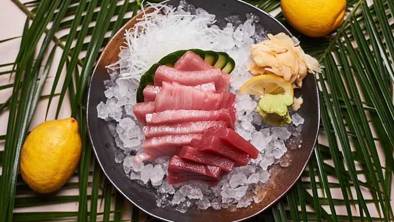 A plate of sashimi slices on ice with shredded daikon, cucumber, pickled ginger, wasabi, a lemon slice, and two whole lemons on a palm leaf background.