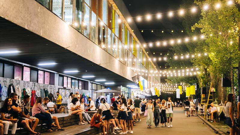 People gather outdoors at night under string lights near a modern building, sitting on benches and walking along a lively pathway.
