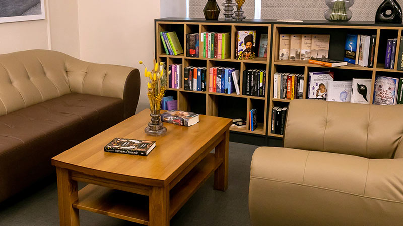 A cozy reading area with two beige sofas, a wooden coffee table holding books and vases, and a bookshelf filled with books and decorative items in the background.