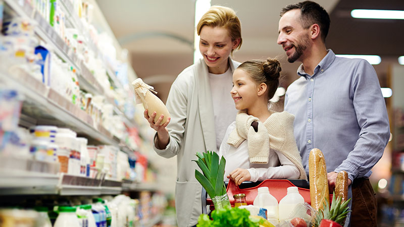 A family of three shops in a grocery store, smiling while looking at a product. The cart is filled with various items including fruits, vegetables, bread, and milk.