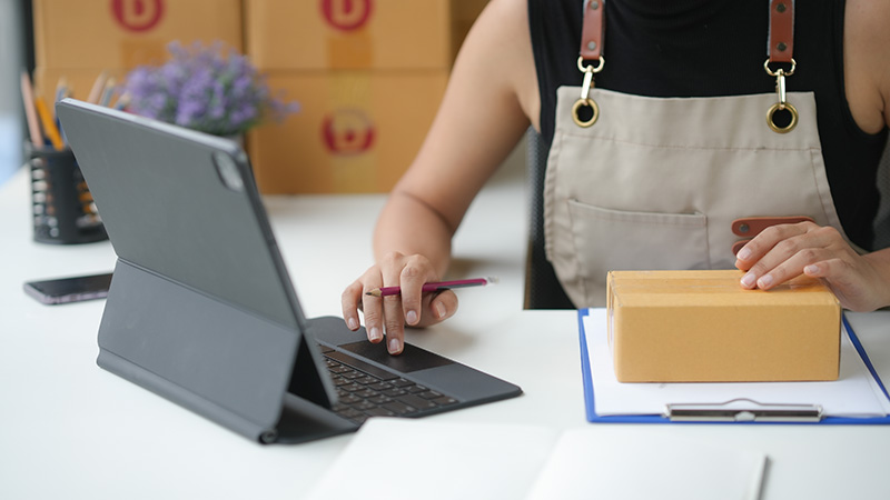 A person wearing an apron works on a tablet with a stylus while holding a small cardboard box over a clipboard on a desk.