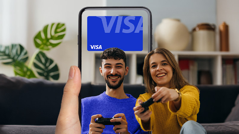 A hand holds a phone displaying a digital Visa card; in the background, a smiling man and woman sit on a couch playing video games.