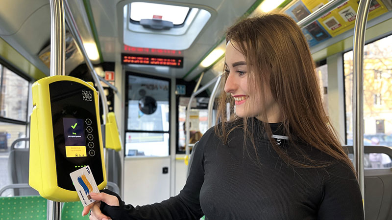 A person with long hair, wearing a black top, uses a card on a yellow ticket machine inside a public bus.