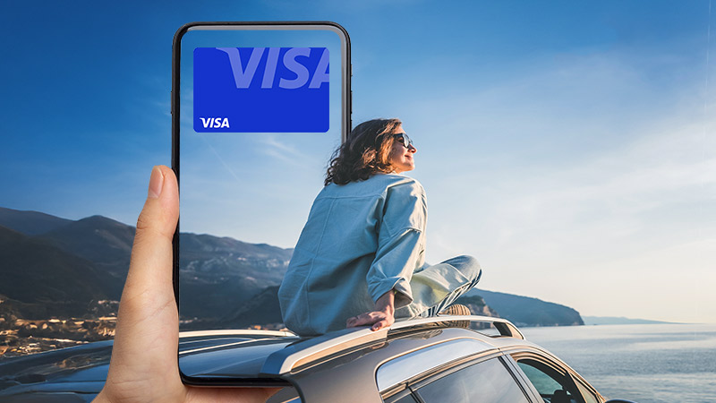 A person sits on top of a car by the ocean, with a hand holding a phone in the foreground displaying a digital Visa card.