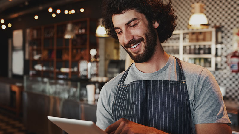 A man with curly hair and a beard, wearing an apron, smiles while using a tablet in a café or restaurant setting with shelves and lights in the background.