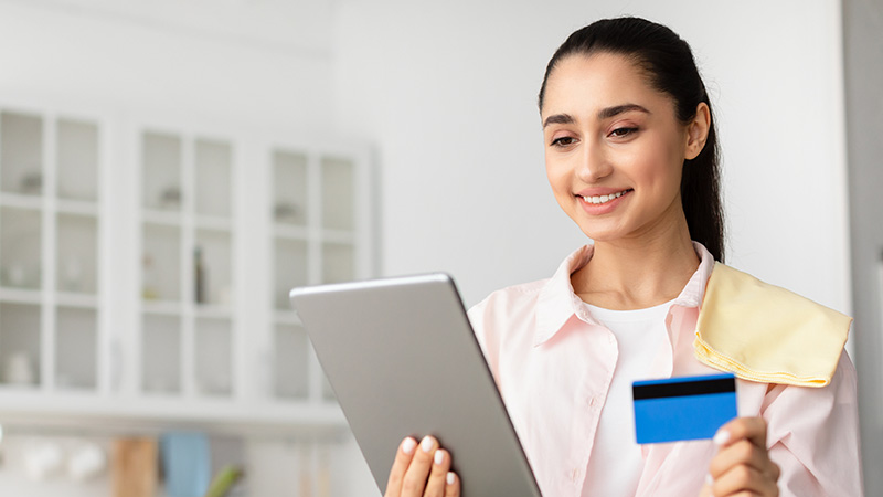 A smiling person holds a tablet and a credit card in a brightly lit kitchen, appearing ready to make an online purchase.